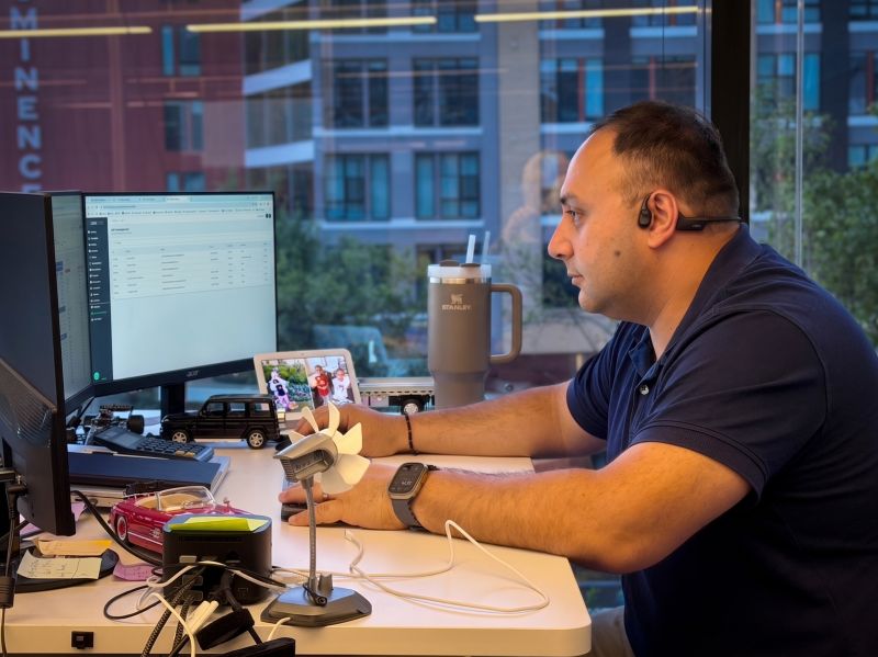 Beka, Carpool Logistics Director of Operations, works at his desk in Carpool's Atlanta office wearing a headset, with a dispatch-style spreadsheet on his monitor, model cars on the desk, and city buildings visible through the window behind him.