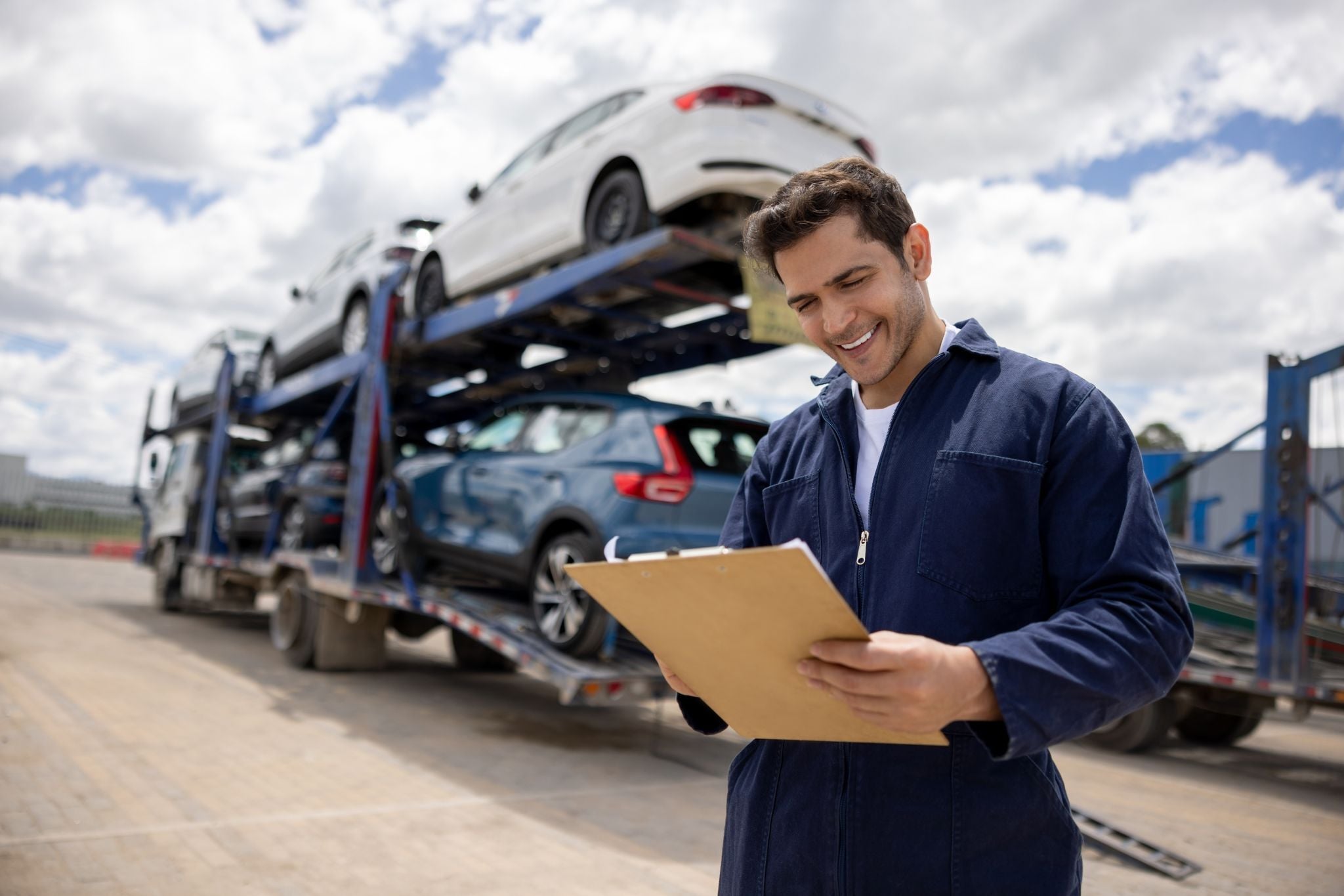 Smiling carrier driver in navy coveralls reviewing paperwork on a clipboard in front of a loaded two-level car hauler.